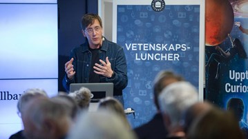 Föreläsare pratar inför publik, rollup med ordet vetenskapsluncher syns i bakgrunden.