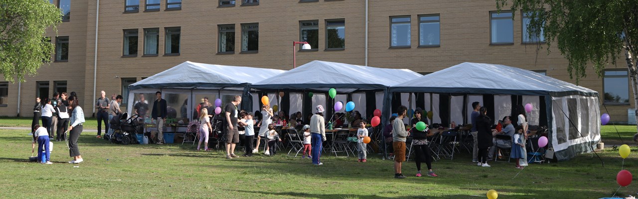 A group of people with baloons standing in front of a building and white tents.