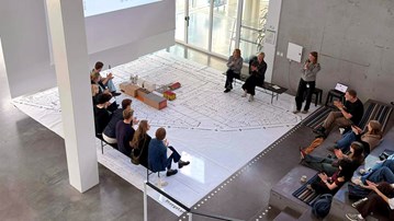 Lecture in big open space with lecturer's, people watching and a big urban plan on the floor