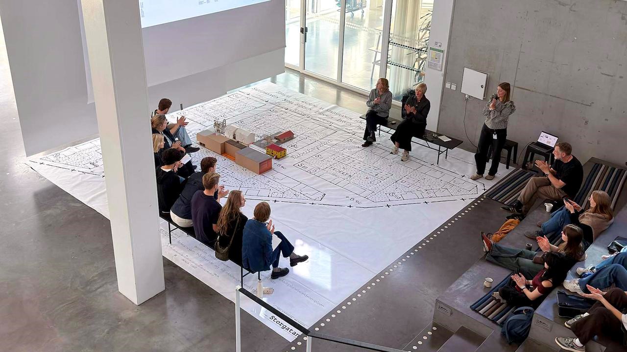 Lecture in big open space with lecturer's, people watching and a big urban plan on the floor