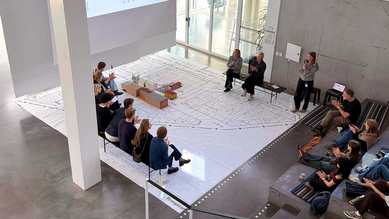 Lecture in big open space with lecturer's, people watching and a big urban plan on the floor
