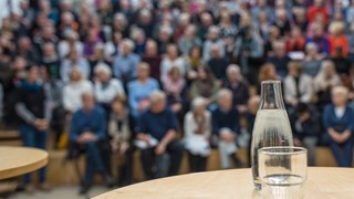 Bild på vattenkaraff med glas på ett bord framför en publik. Bilden ska illustrera att hålla föredrag.