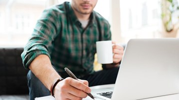 Person sitter i en soffa och arbetar. Har dator, kaffekopp och block där han skriver.