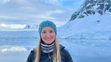 Kylie Owen står i mörkblå vinterjacka och ljusblå mössa och ler mot kameran. I bakgrunden syns en spegelblank sjö, snö och ett berg.