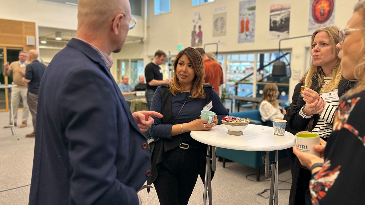 Fyra personer står samlade kring ett bord och pratar med människor minglande i bakgrunden.