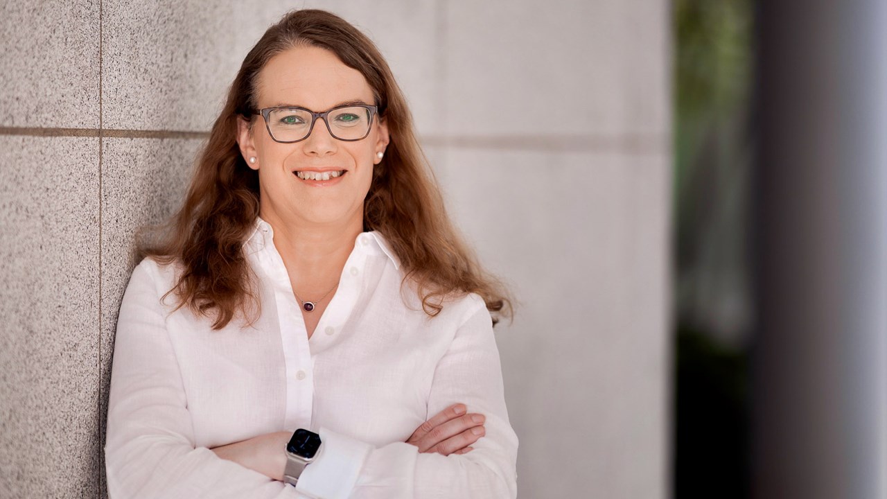 Foto av Alexandra Kafka Larsson leende, med armarna i kors, lutad mot en grå vägg