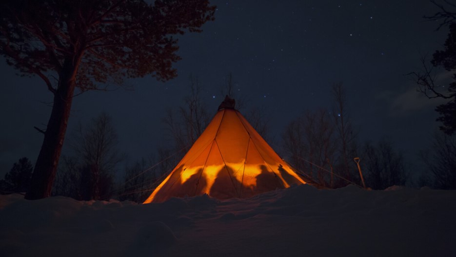 Indigenous Sámi tent with several people sitting in it, lit up by a fire in a dark winter landscape.