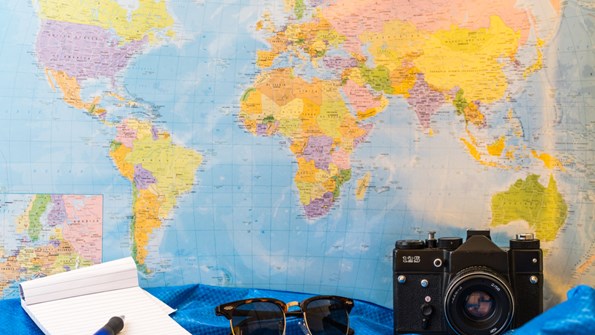 World map in background. Note book, sunglasses and camera in foreground.