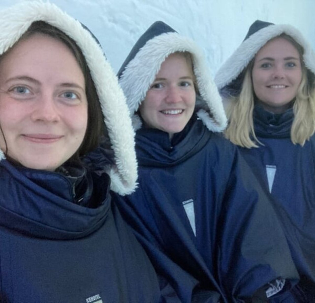 Three young women sitting next to each other in thick coats with fur collars in the ice hotel and looking at the camera and smiling