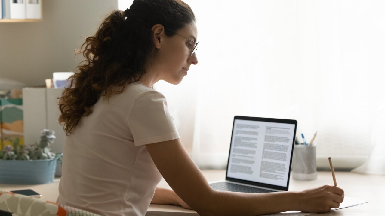 En kvinna sitter vid ett bord med laptop och headset, böcker och antecknar i ett block.