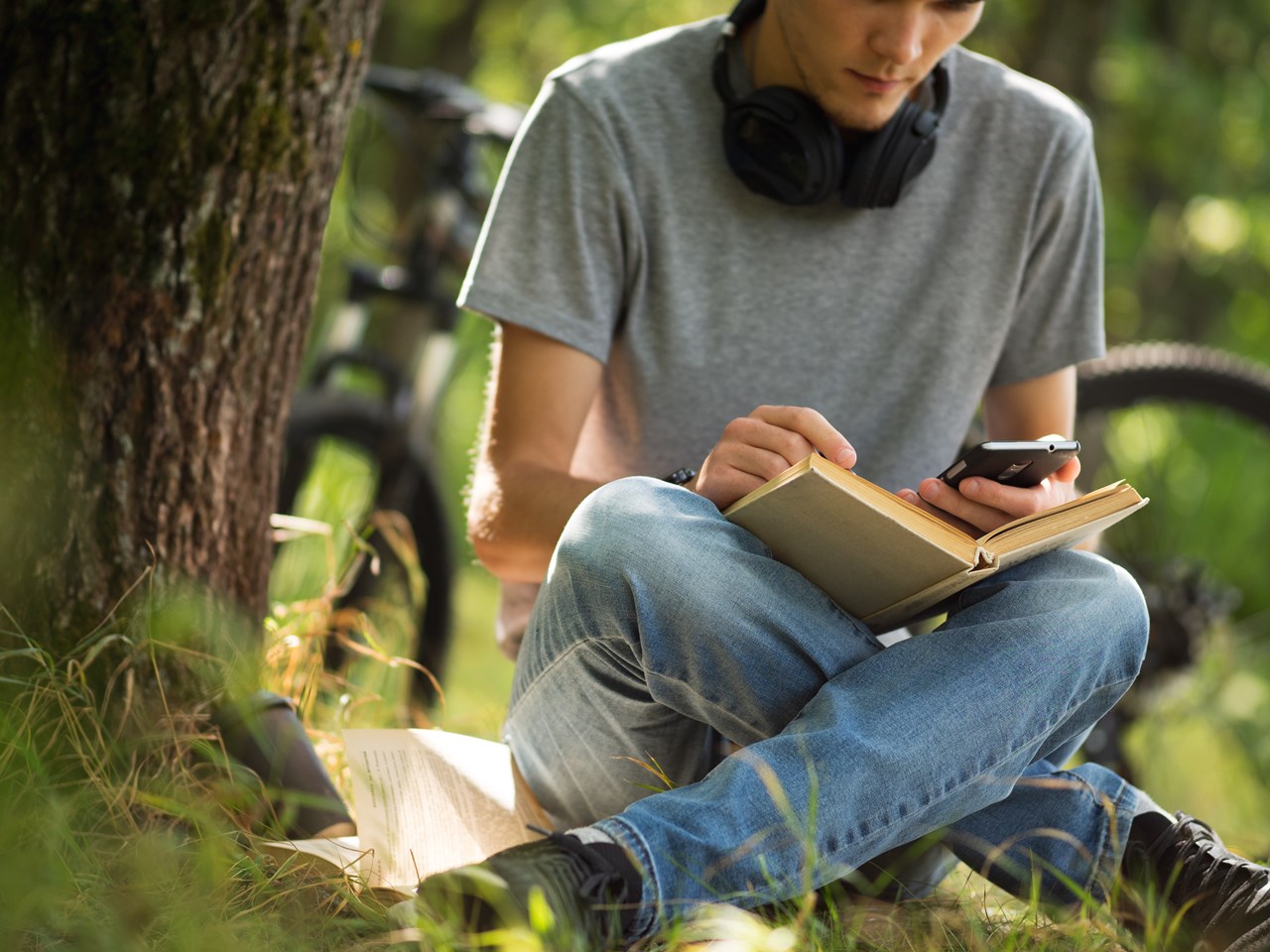 Kille som sitter utomhus i gräset vid ett träd med en mobil och en bok.