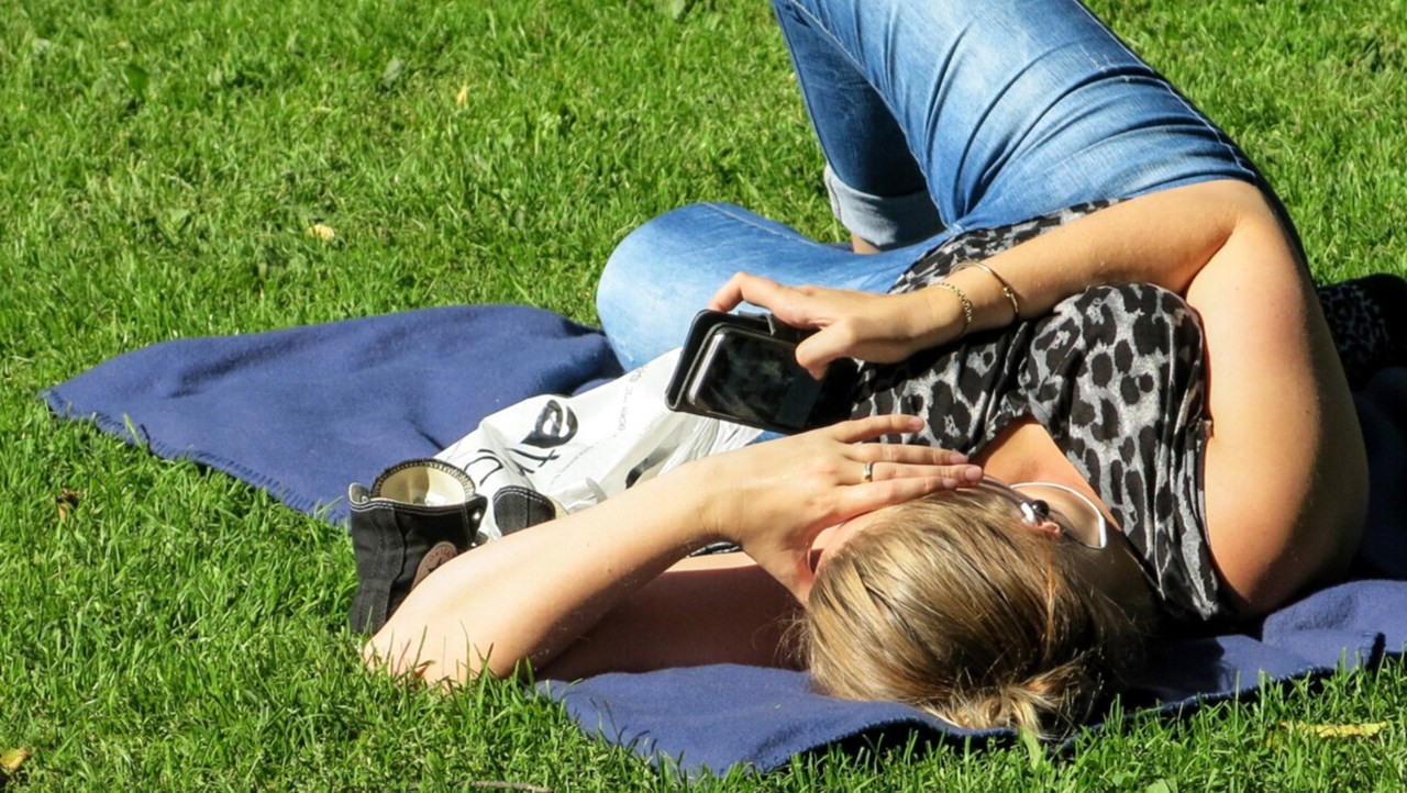 Kvinna med mobiltelefon på ligger på filt på gräsmatta och surfar.