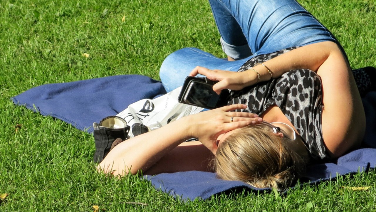 Kvinna med mobiltelefon på ligger på filt på gräsmatta och surfar.