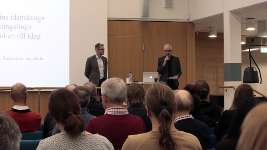 Två män talar inför en sittande publik i Humanisthuset, Umeå universitet
