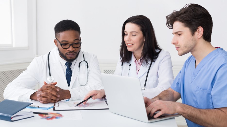 Bilden visar tre personer med medicinrock och medicinsk utrustning, en laptop samt en bok som sitter vid ett bord och samtalar.