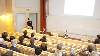 a person is talking in front of an auditorium populated by people. Many of them are wearing suits or equivalent attire.