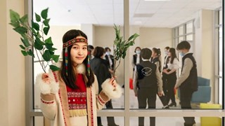 Elev iklädd traditionella kläder som håller upp en lövkvist. Genom fönstren bakom syns fler elever i lärare samlande i ring