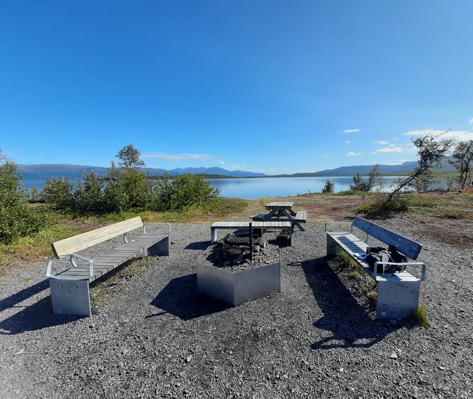 En eldstad nära en sjö i Abisko, med blå himmel och berg i bakgrunden.