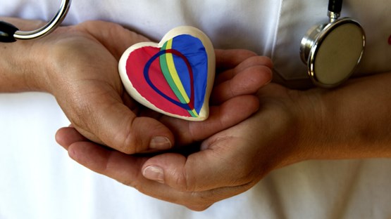 A person in a healthcare worker outfit holding a small heart-shaped pillow with the Sámi flag on it. 