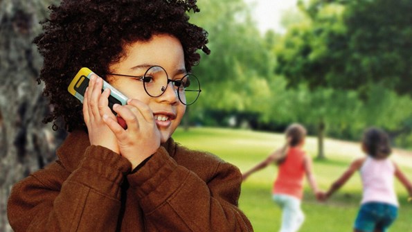 Child speaking on a mobile phone, outside, a couple of other children are playing in the background holding hands