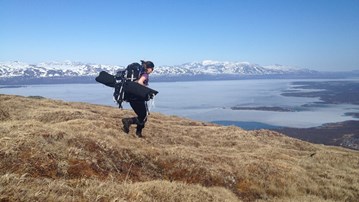 A researcher carrying equipment in a lake and mountain environment