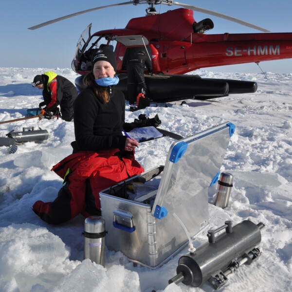 Två personer sitter på is och tar prover med en helikopter i bakgrunden