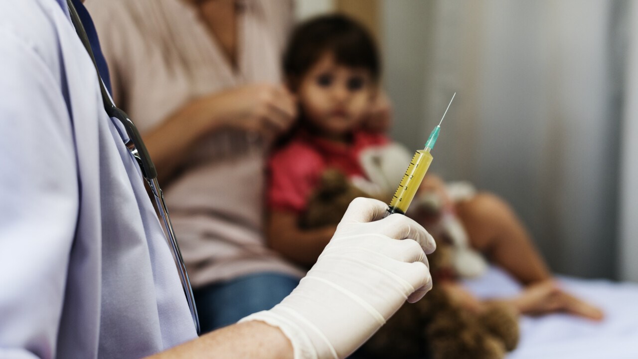 Närbild på sjukvårdspersonal med spruta i hand, redo att vaccinera ett barn i bakgrunden.