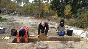 Three persons kneeling in fron of a digging trench in a forest area