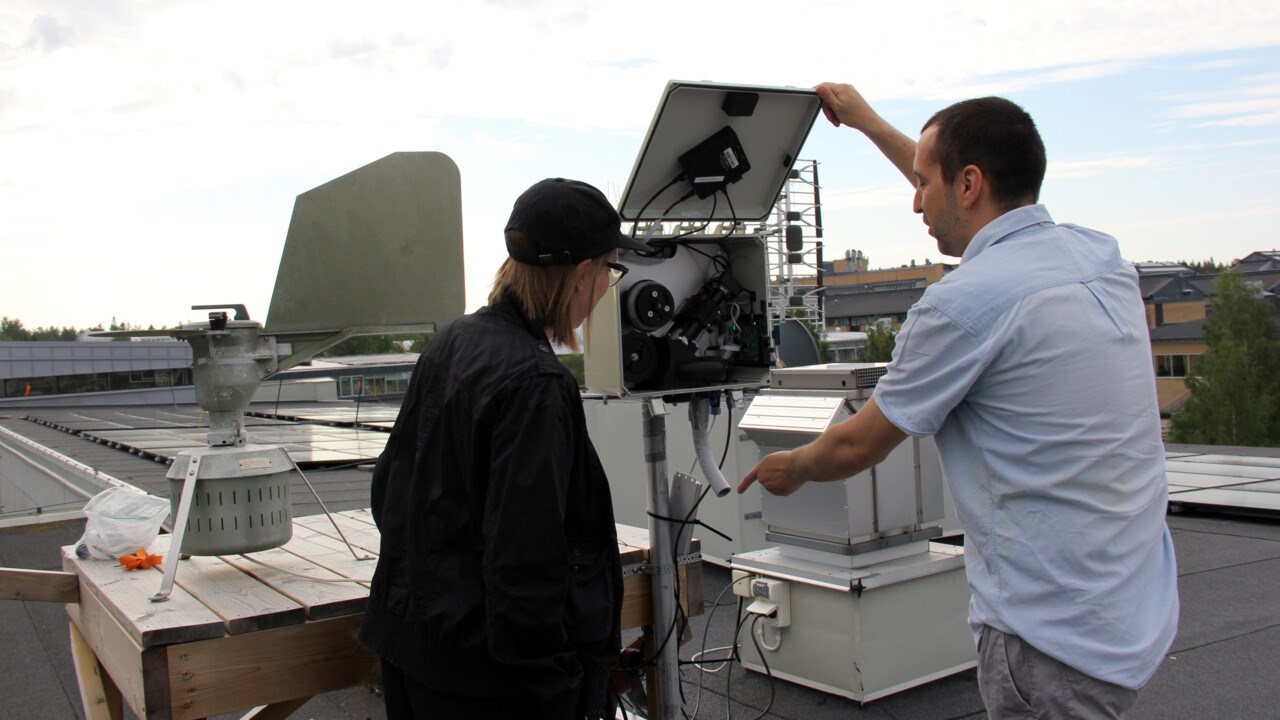 Två personer står på ett tak framför en elektronisk apparat med ett rörformat munstycke