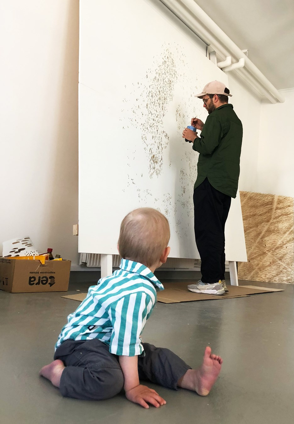 A man is painting with tar on a canvas, and a toddler is watching in the foreground