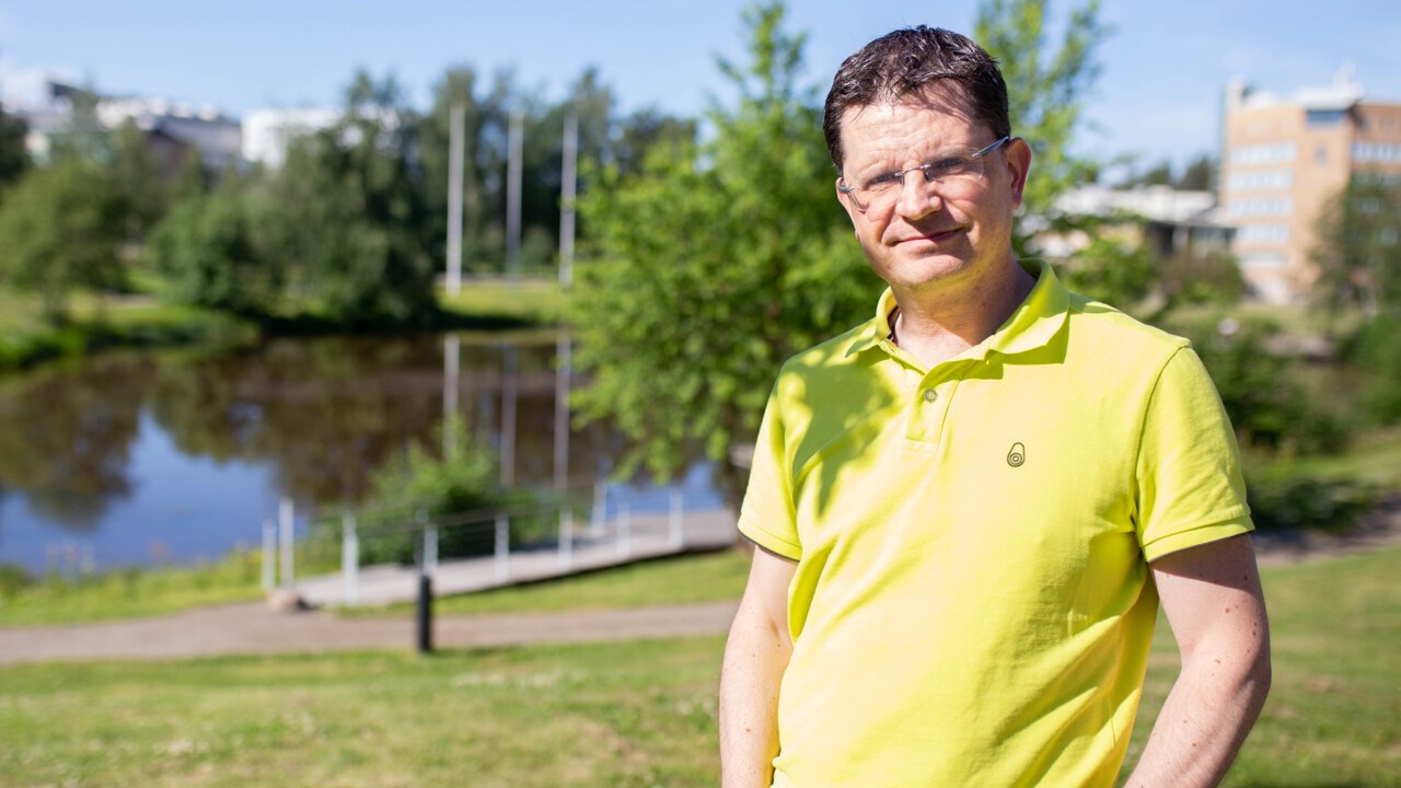 Medelålders man i limegul tröja placerad på ett soligt campus med dammen i bakgrunden.