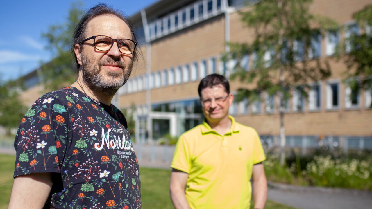 Två medelålders män, den ena i knallgul t-shirt och den andra i mönstrad t-shirt. Står på ett soligt campus.