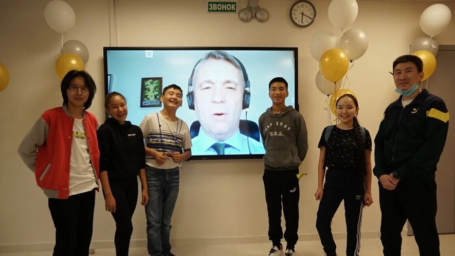 Elever står på vardera sida om en storbildsskärm om ler mot kameran medan Peter Skölds välönskningar syns på skärmen i bakgrunden