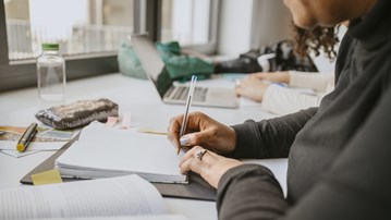 En person som skriver med penna och papper och en person som sitter och arbetar vid laptop. 