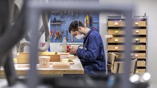 A young man in a face mask is bent over a table, sanding a model. A tool board with chisels, pliers and rasps is visible in the background.