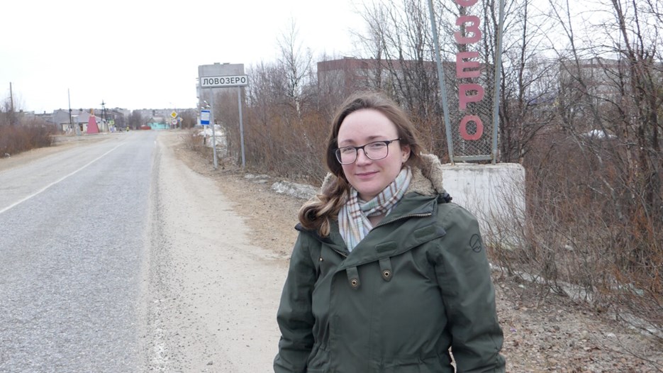 Woman stands by road, in front of roadsigns with Cyrillic text.