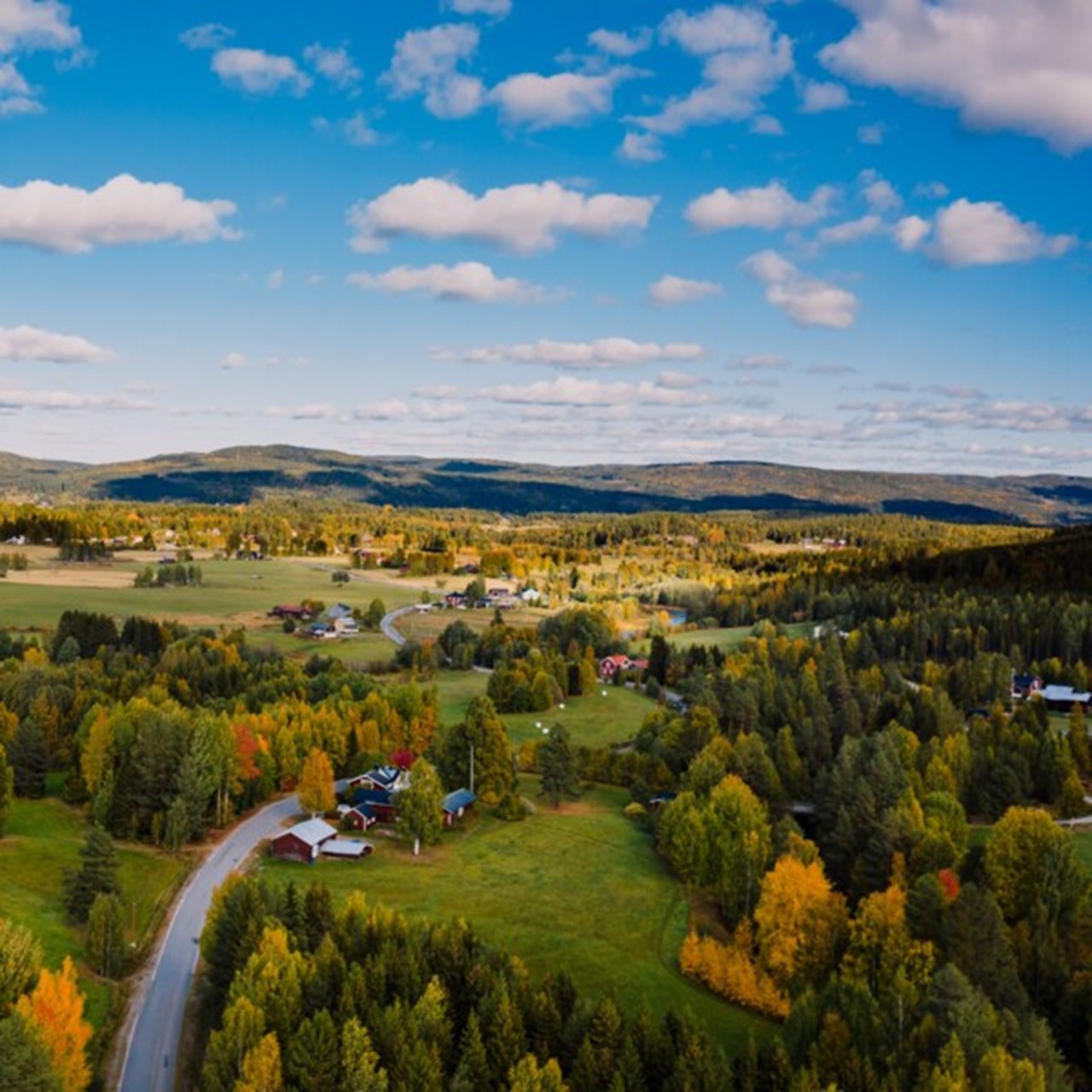 ser en väg som slingrar sig genom en by med jordbrukslandskap om hösten. I bakgrunden syns bergskullar. Himlen är blå med fluffiga moln