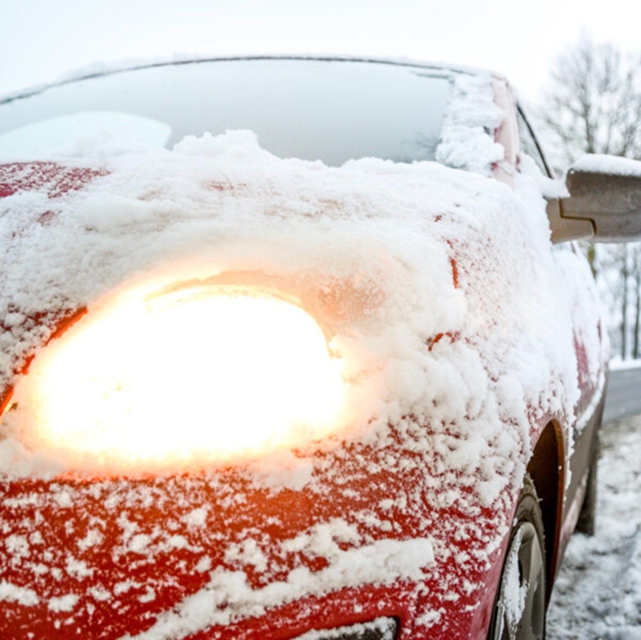 he headlight is on and shines through the ice of a snow-covered red car