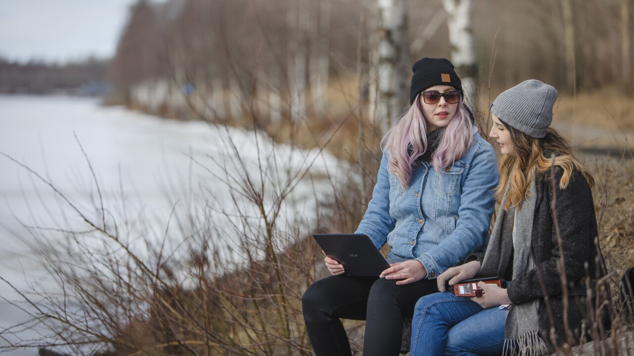 Studenterna Evelinn Carlsson och Tinah Ekwall sitter vid älvskanten med en laptop i knäet..