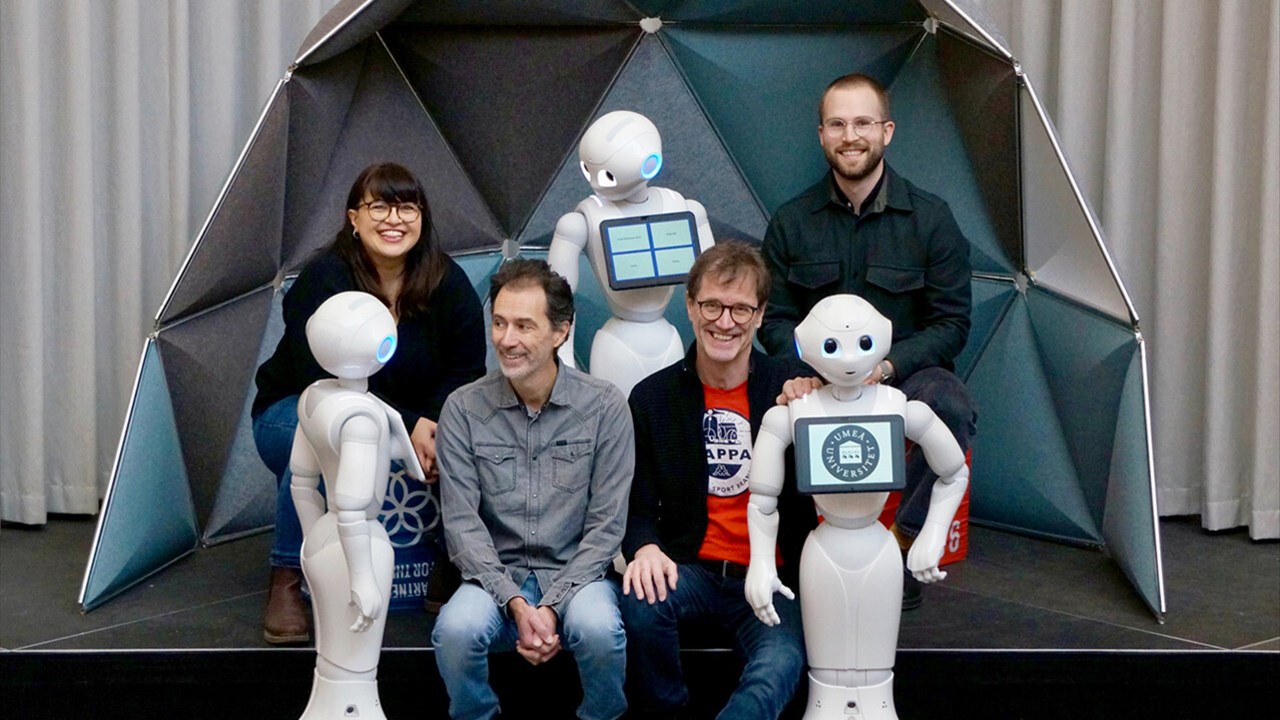 Suna Bensch, universitetslektor i datavetenskap, Filip Edström, doktorand på Handelshögskolan, Xavier de Luna, professor vid Handelshögskolan, och Thomas Hellström, professor i robotik, sitter med tre robotar som tittar på dem.