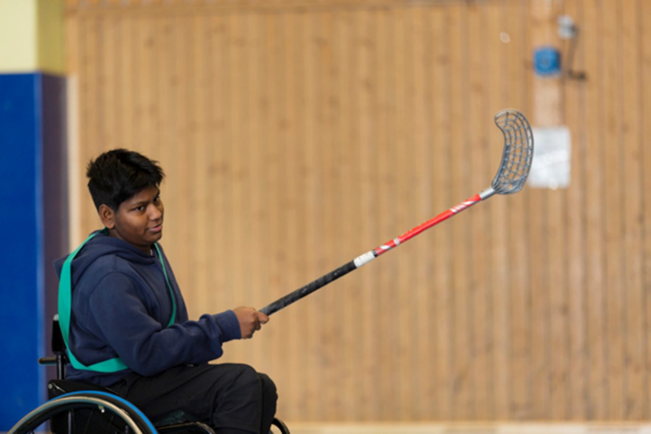 Kille i rullstol med innebandyklubba i handen skjuter iväg en boll.