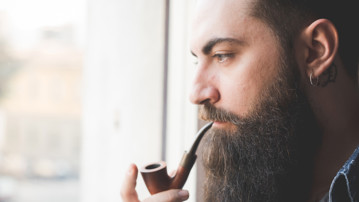 Young bearded man smoking pipe by window