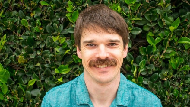 A man, wearing a blue shirt, with a rich moustache and smile on hisface is standing in front of a thick, bright green hedge.