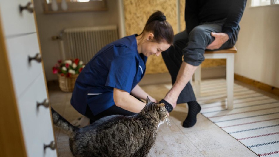 Kvinna i hemtjänsten som hjälper en person på med strumporna. En katt sysns i förgrunden.