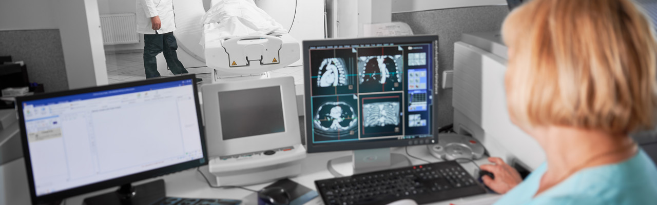 Medical computed tomography or MRI scanner. Back view of blonde nurse sitting at computer, doctor standing, making MRI. Specialists working in clinic. Concept of modern diagnostics.