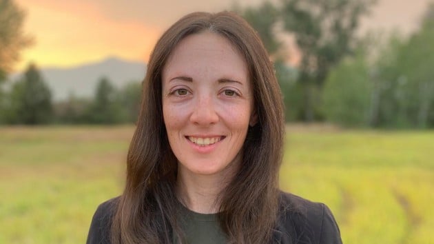 A woman with long dark hair, wearing dark casually elegant clothes, isstanding in an open field during a colourful sunset.