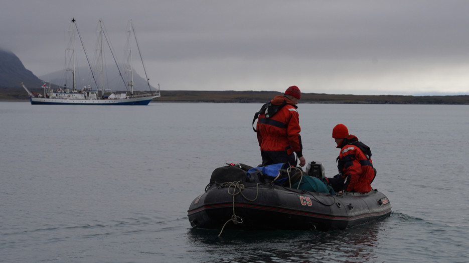 Two people on a small boat, and a bigger boat in the background.