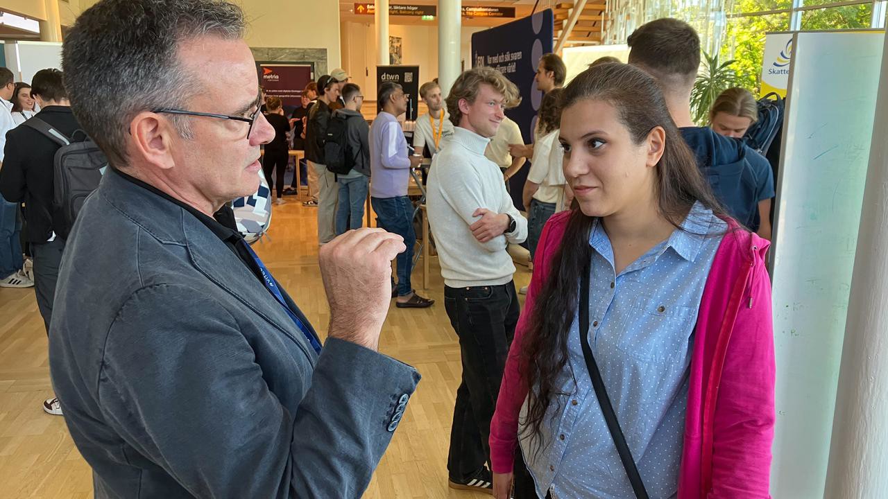 Bild på Anders Sjövik och Simona Ebid i samtal under mingel bland företagsmontrar.