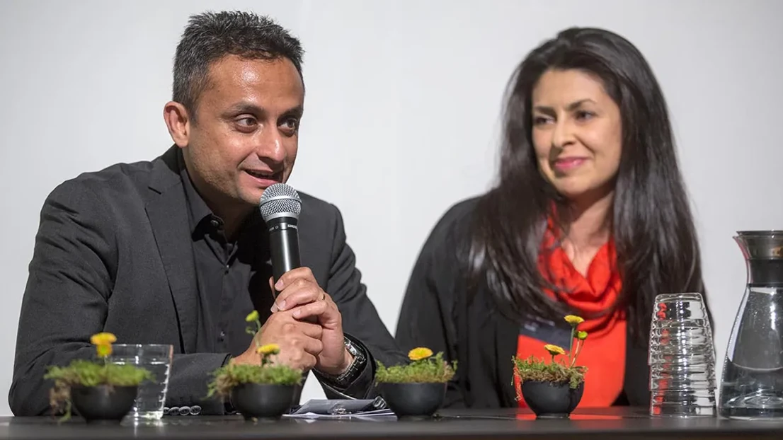 One man and a woman sitting at a table. The man is talking in to a microphone