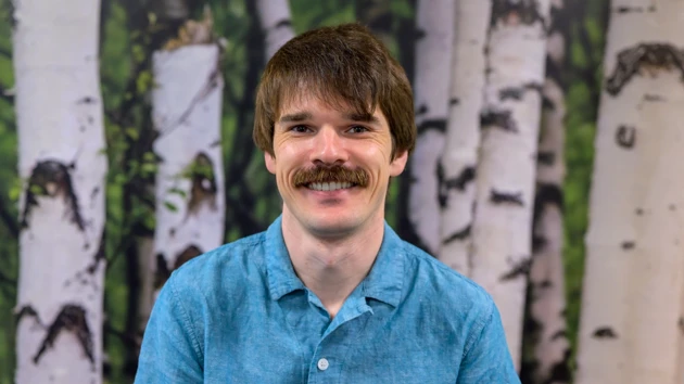 A man with a mustache and a blue shirt poses in front of a birch tree background